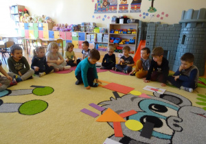 Chłopiec układa pajaca z figur geometrycznych według wzoru, dzieci siedzą wokół niego i obserwują z zainteresowaniem.